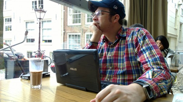 Sascha im Cafe in Amsterdam. Stilsicher mit Schirmmütze und Karohemd, vor ihm sein Asus-Laptop und ein Hipsterkaffee.